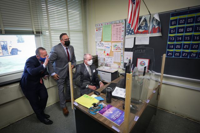 Governor Phil Murphy sitting at a teacher's desk in the corner of a classroom that is partitioned by plastic. US Education Secretary Miguel Cardona is standing behind him.
